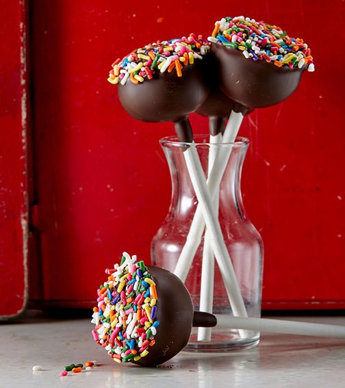 3 chocolate dipped cake pops with rainbow sprinkles in a small glass jar. 1 cake pop on the table in front. Red background