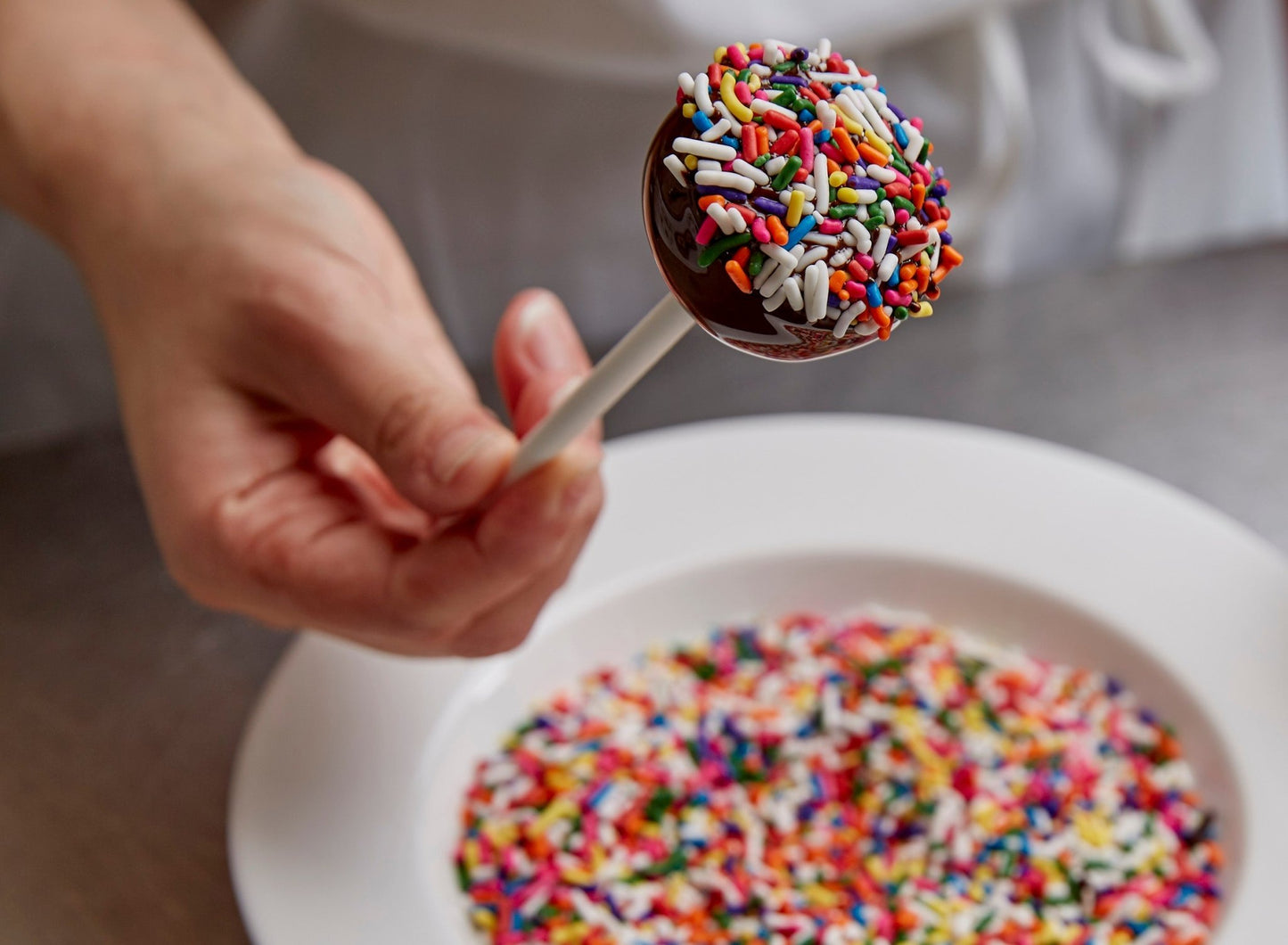 
                  
                    A close up of a check dunking chocolate covered cake pops into a bowl of rainbow sprinkles
                  
                