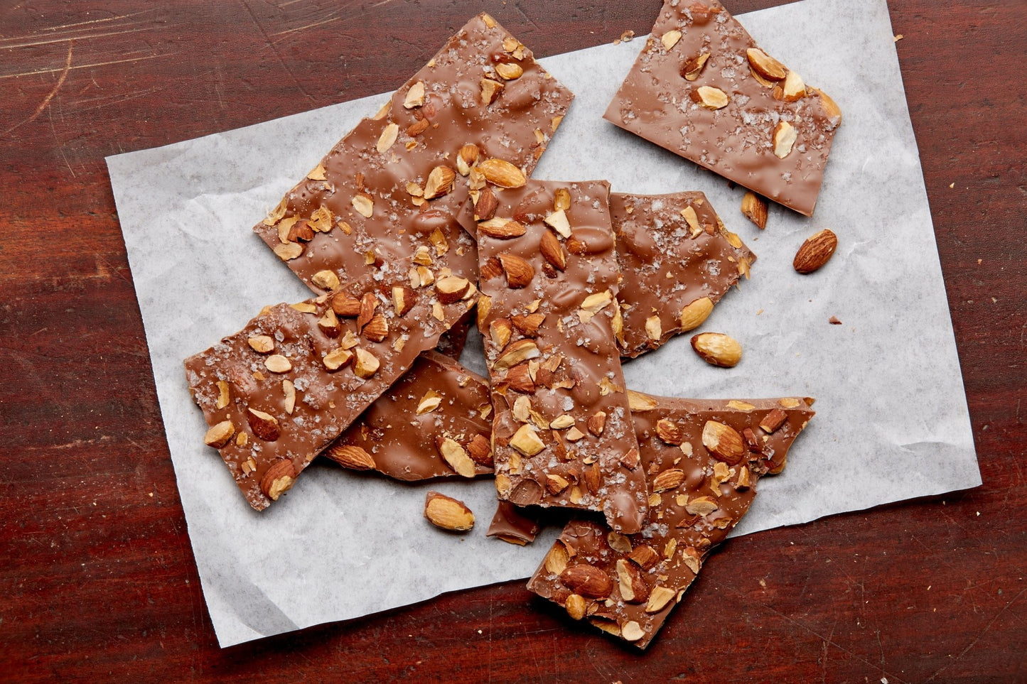 
                  
                    Large pieces of milk chocolate bark with almonds and sea salt on parchment paper on a rustic wooden tabletop
                  
                