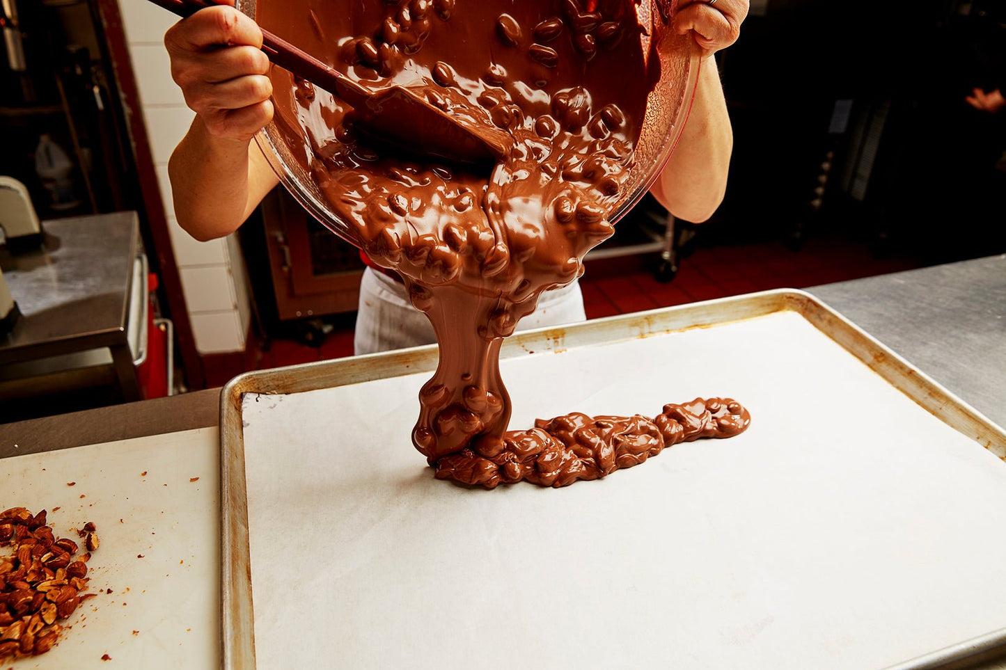 
                  
                    Chef in the process of making milk chocolate bark pouring chocolate mixed almonds onto a parchment paper line sheet tray
                  
                