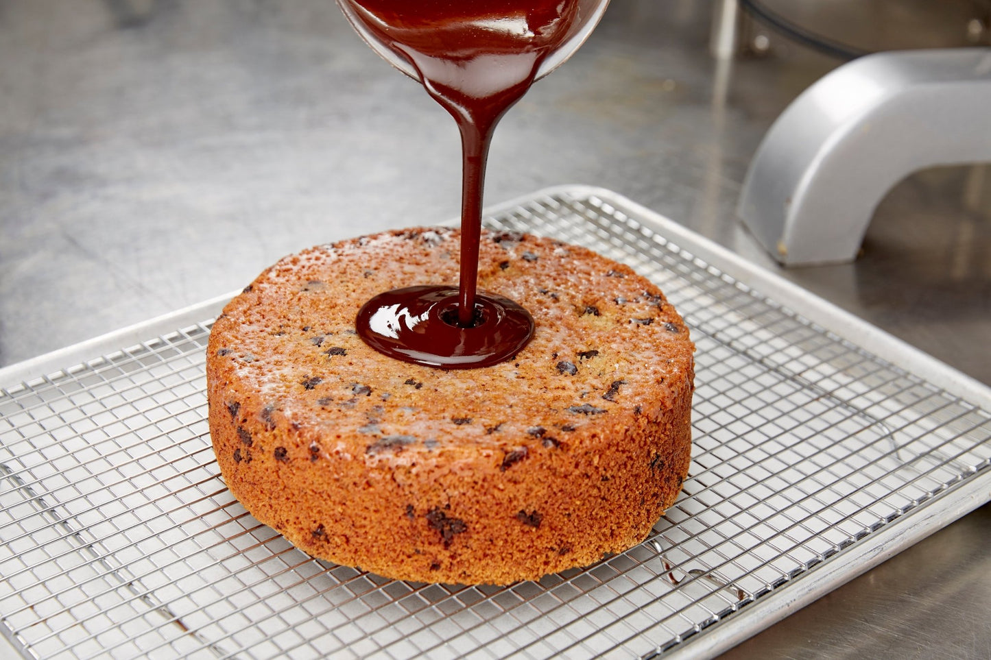 
                  
                    Chocolate being poured on a chocolate almond cake. Cake is a sheet tray with a rack on it. Tray is on a stainless table
                  
                