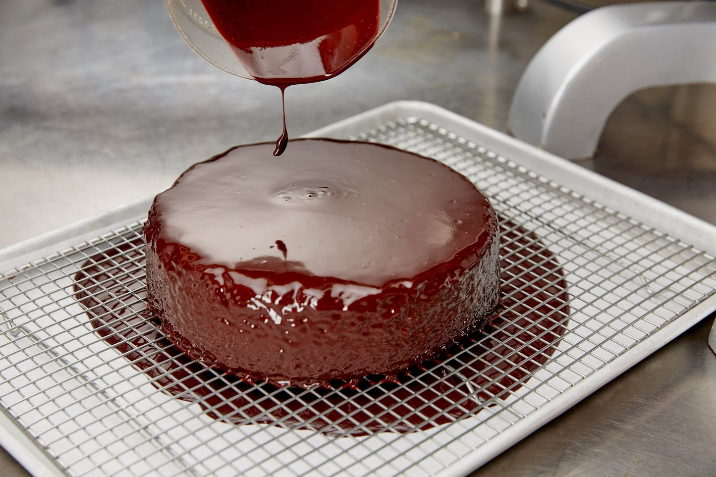 
                  
                    Chocolate being poured on a chocolate almond cake. Cake is a sheet tray with a rack on it. Tray is on a stainless table
                  
                
