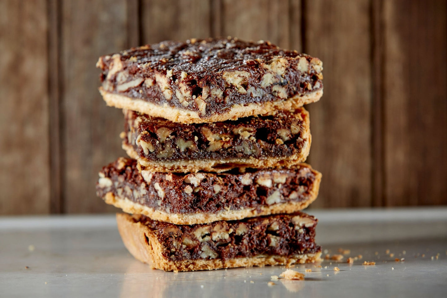 A close up of 4 chocolate pecan bars with crumbs on the tabletop in front of a rustic wooden paneled background