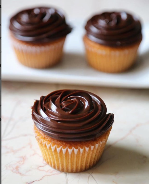 3 chocolate frosted cupcakes, 1  in foreground on a marble table, 2 blurred on a plate in the background