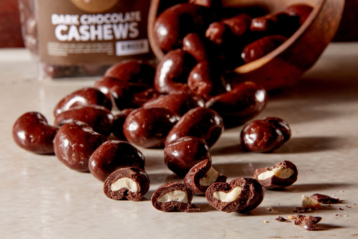 Dark chocolate cashews poured from a wooden bowl some broken open in the front and a branded bag of them in the background