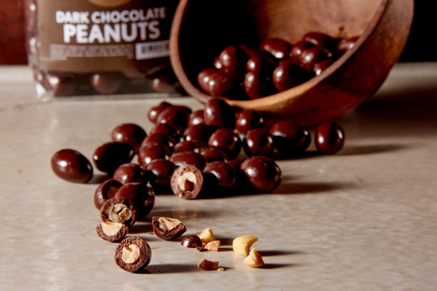 Dark chocolate peanuts poured from a wooden bowl some broken open in the front and a branded bag of them in the background