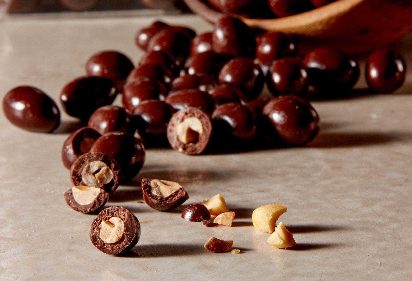 Close up of chocolate covered peanuts poured from a wooden bowl with a few broken open in the front