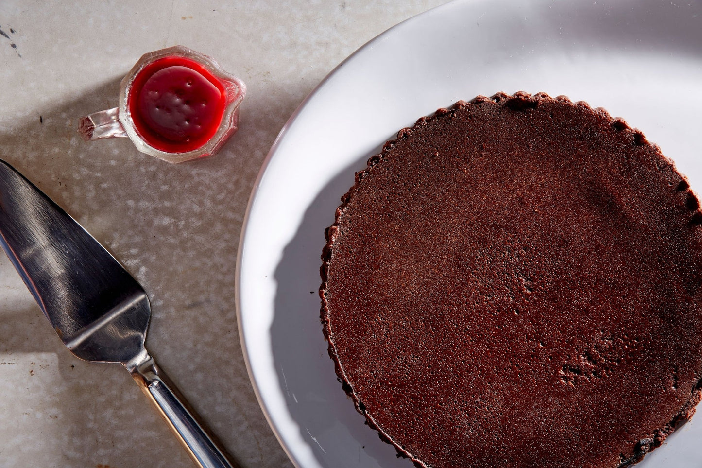 Overhead whole gluten free Flourless Chocolate Cake on a white plate next to a raspberry sauce boat and a silver cake slicer