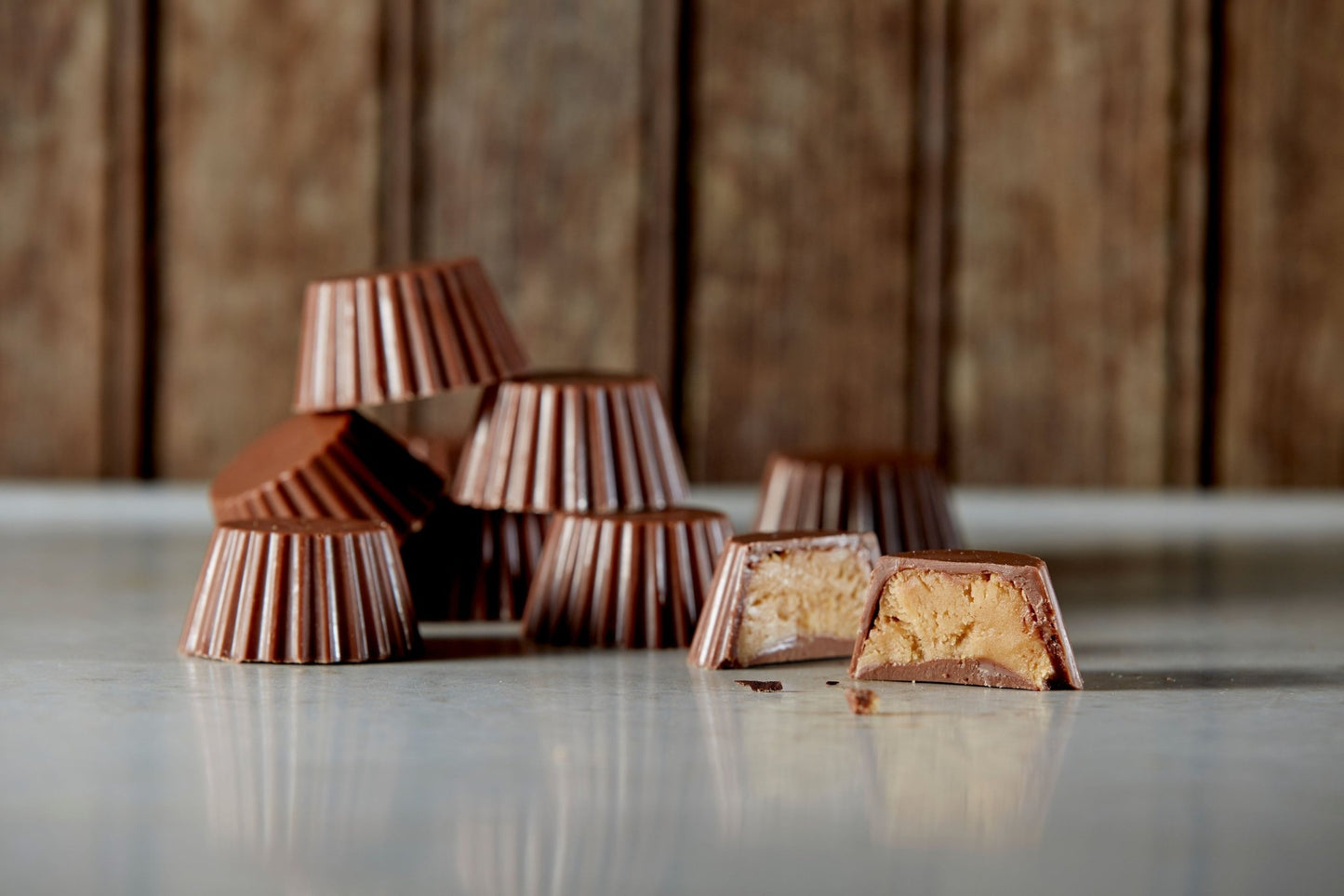 A pile of milk chocolate peanut butter cups with 1 cut open. They are on a vintage tabletop in front of an aged panned wall