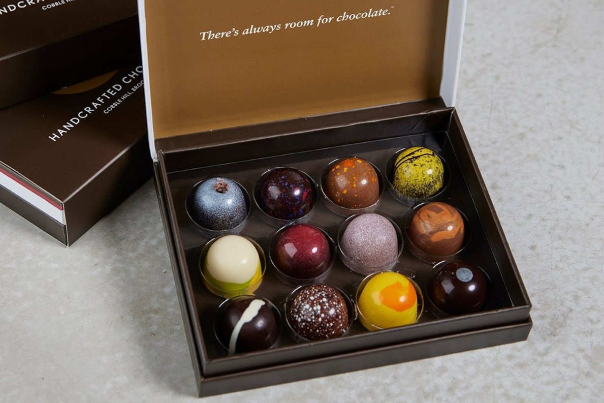 Open box of 12 handcrafted, colorful bonbons alongside branded boxes - inside says There is always room of chocolate
