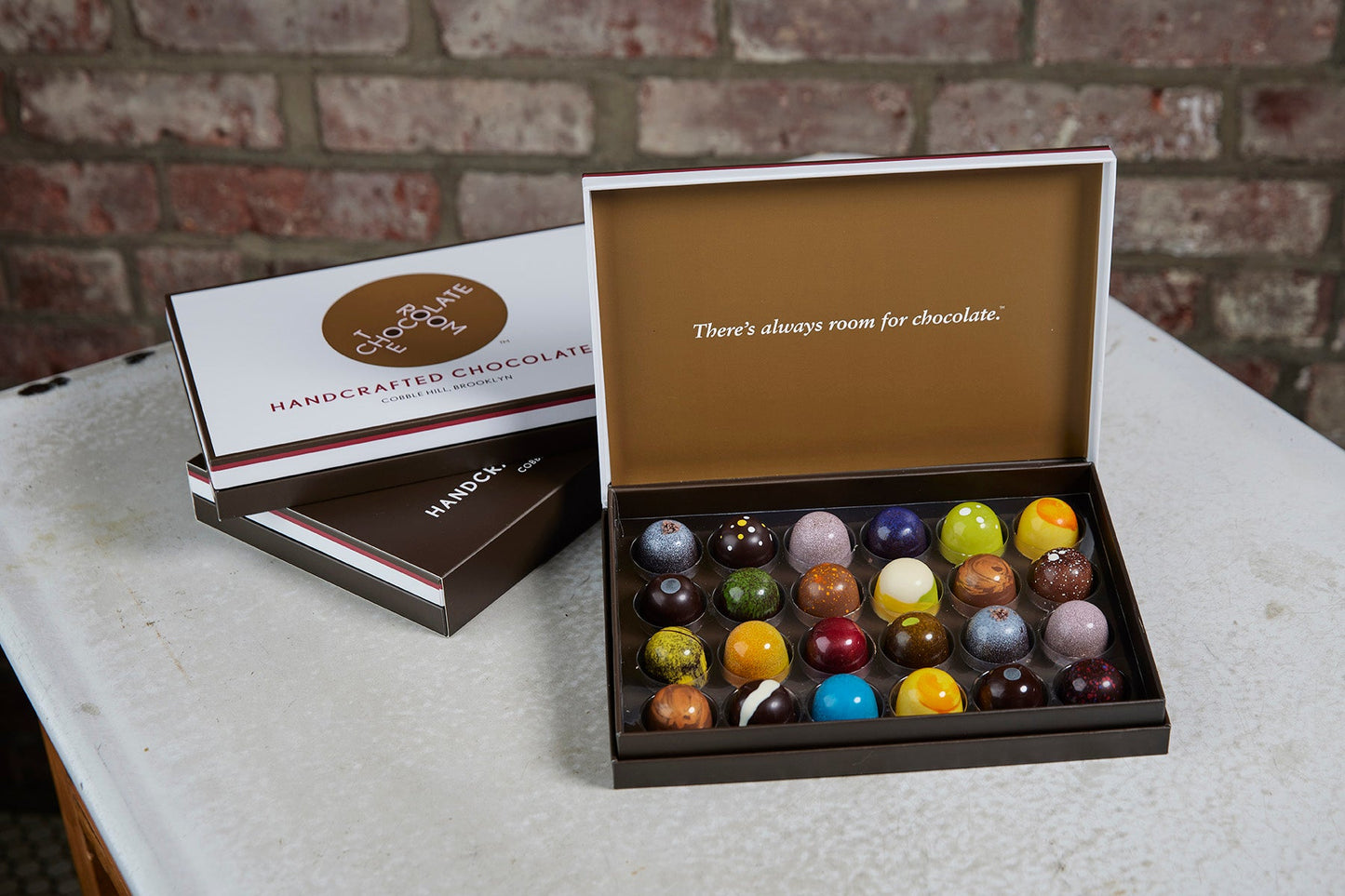 Open box of 24 handcrafted, colorful bonbons alongside branded boxes - inside says There is always room of chocolate