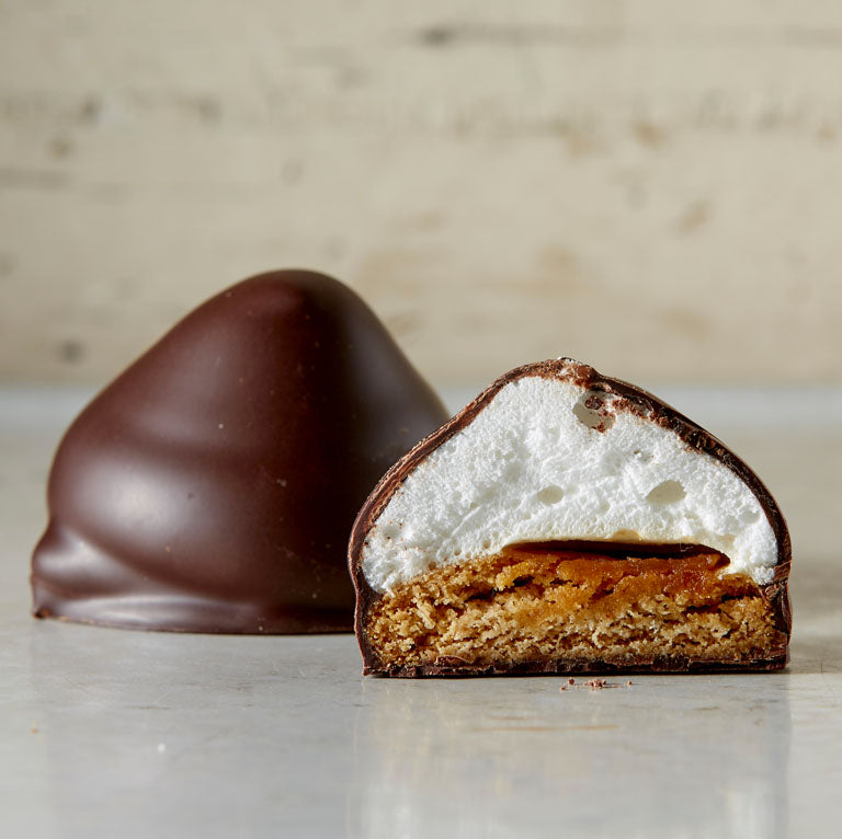 Close up of 2 dark chocolate s'mores, 1 whole, 1 cut in half revealing the graham cracker and domed marshmallow, with crumbs 