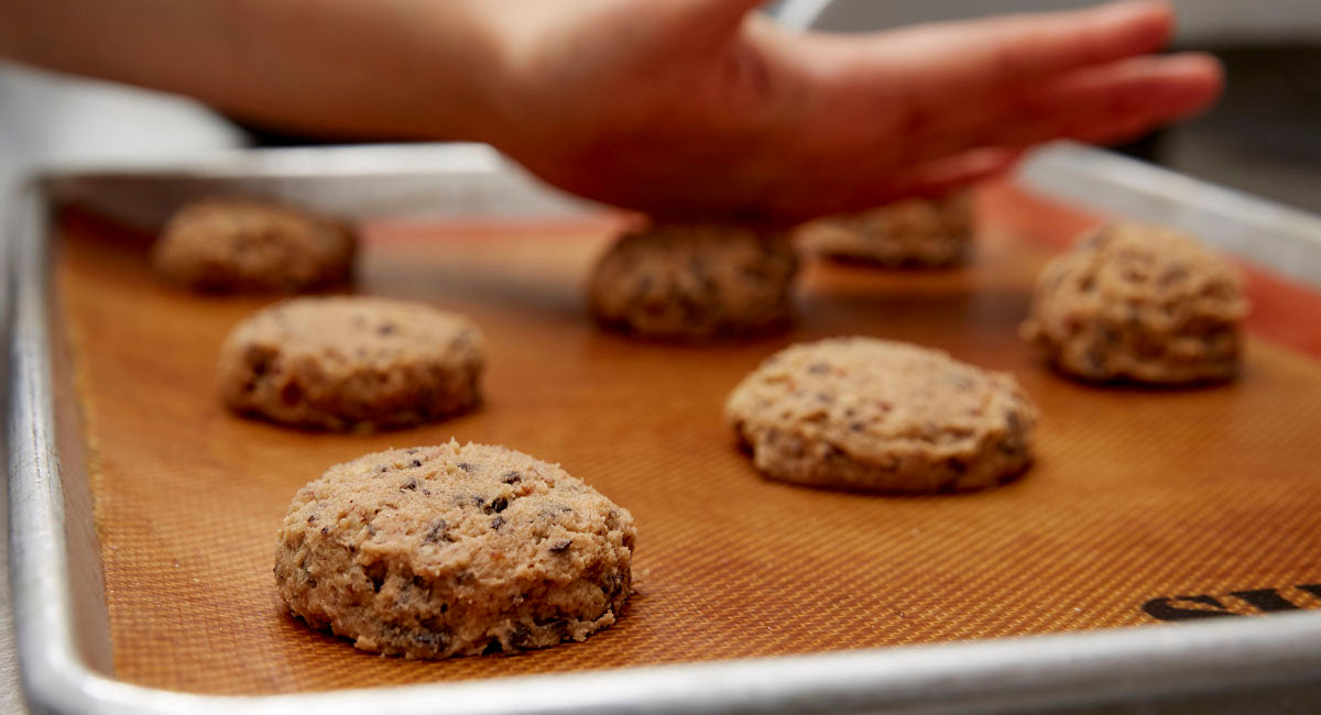 Portioned cookie dough on a sheet tray being pressed down by chef prior to being baked