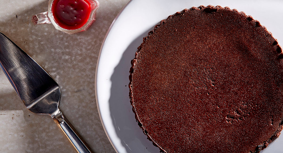 Overhead whole gluten free Flourless Chocolate Cake on a white plate next to a raspberry sauce boat and a silver cake slicer
