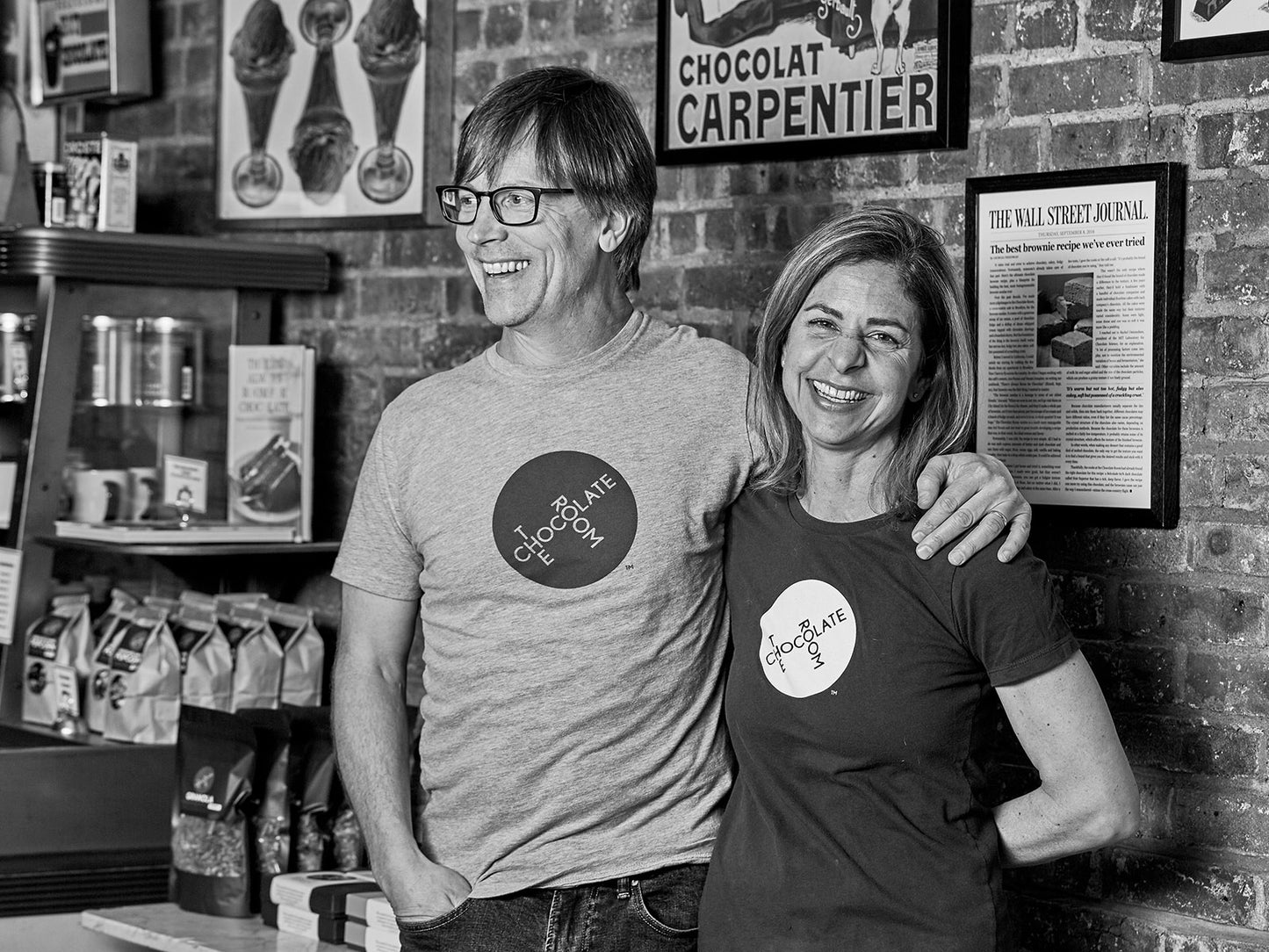 A happy Jon and Naomi, The Chocolate Room founders, in front a decorated brick wall in the shop