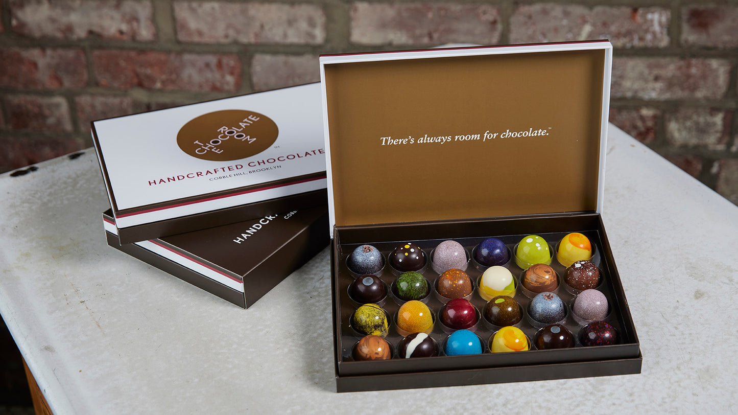 Open box of24  handcrafted, colorful bonbons alongside branded boxes - inside says There is always room of chocolate