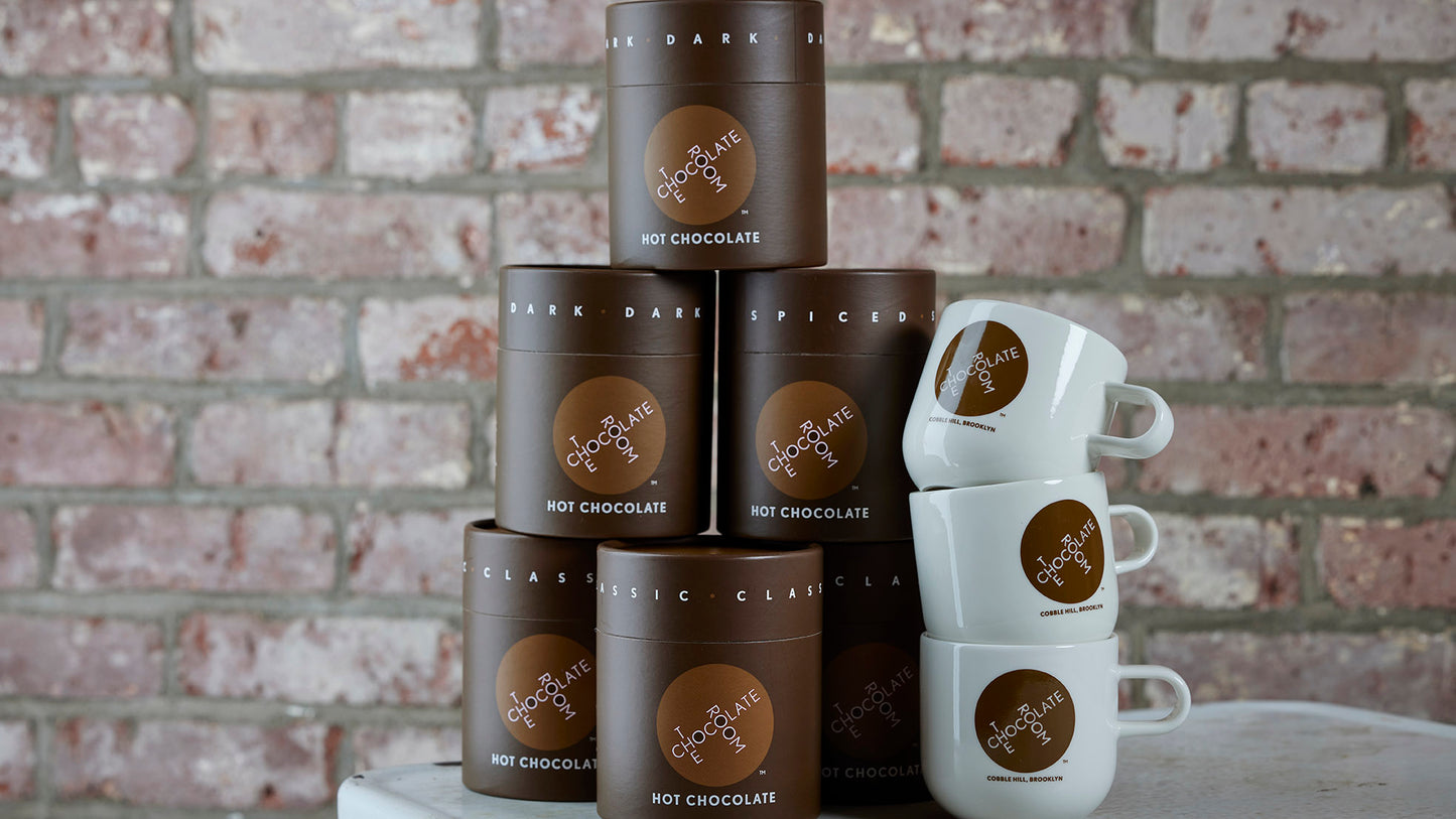 A stack branded hot chocolate powder tubes next to a stack of branded mugs